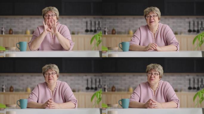 可爱的老年妇女坐在厨房里看着相机，说话和挥手欢迎，在线视频聊天概念