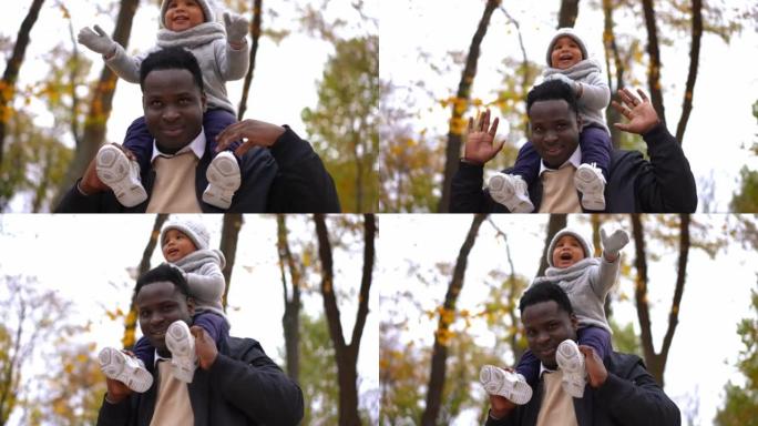 快乐的非洲裔美国年轻人看着相机微笑的肖像，迷人可爱的男孩坐在肩膀上。自信的父亲在秋天公园的户外摆姿势