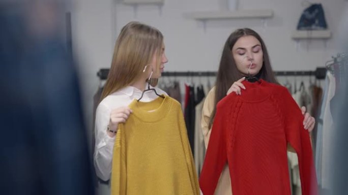 两个年轻漂亮的女人在黑色星期五的商店里选择红色和黄色的毛衣，改变衣架，欣赏镜子里的反射。积极的白人购