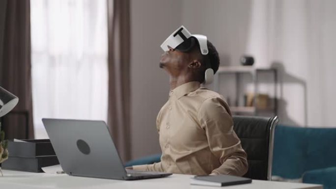 使用虚拟现实进行教育，年轻的黑人戴着头戴式显示器，坐在笔记本电脑的桌子旁，环顾四周