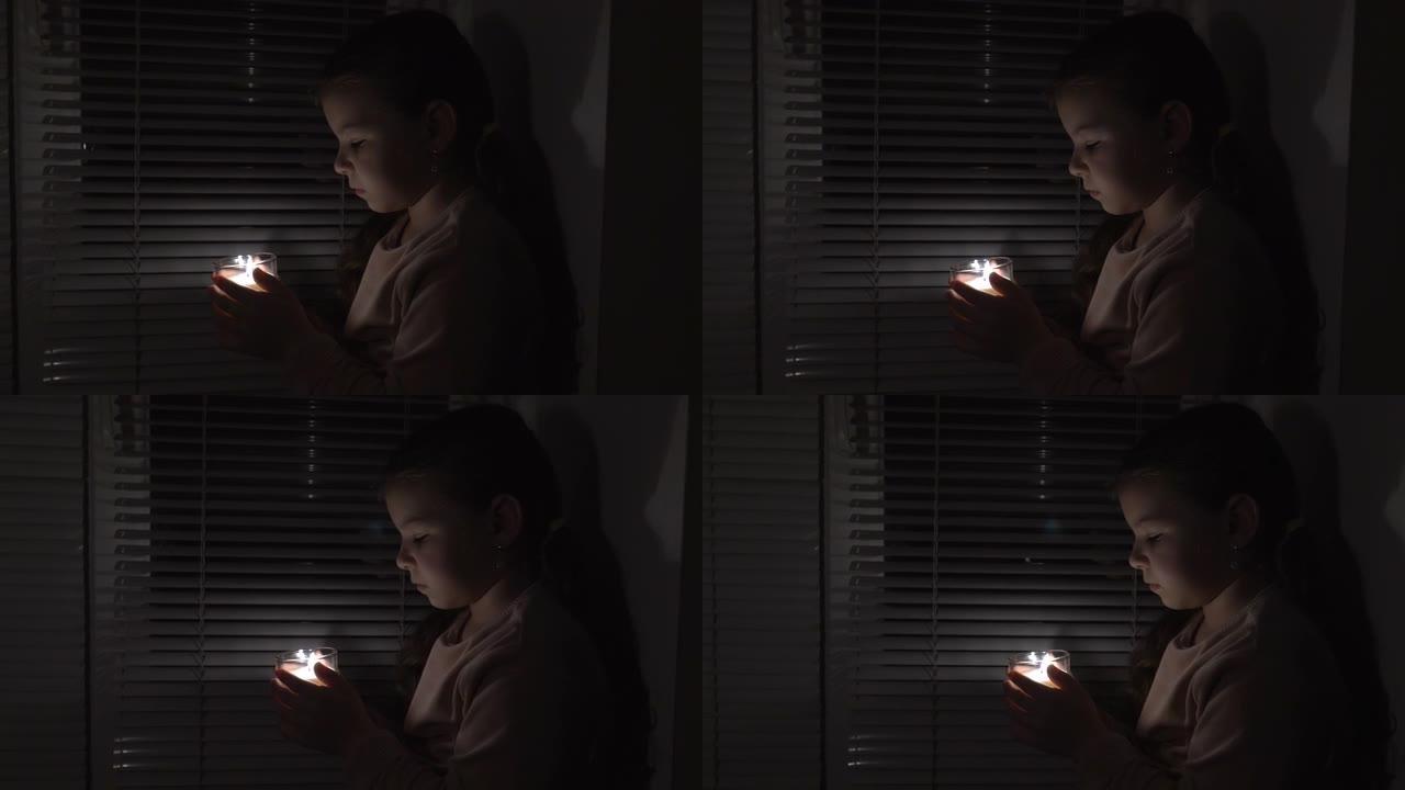 一个女孩在窗边的黑暗房间里，手里拿着玻璃杯里的蜡烛