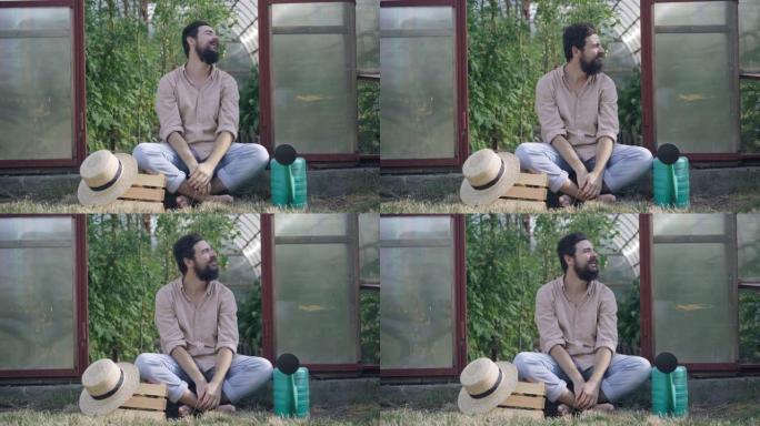心满意足快乐微笑的男农夫坐在温室门口伸伸懒腰四处张望。宽镜头的黑发白人留着胡子的男人休息在收获蔬菜。