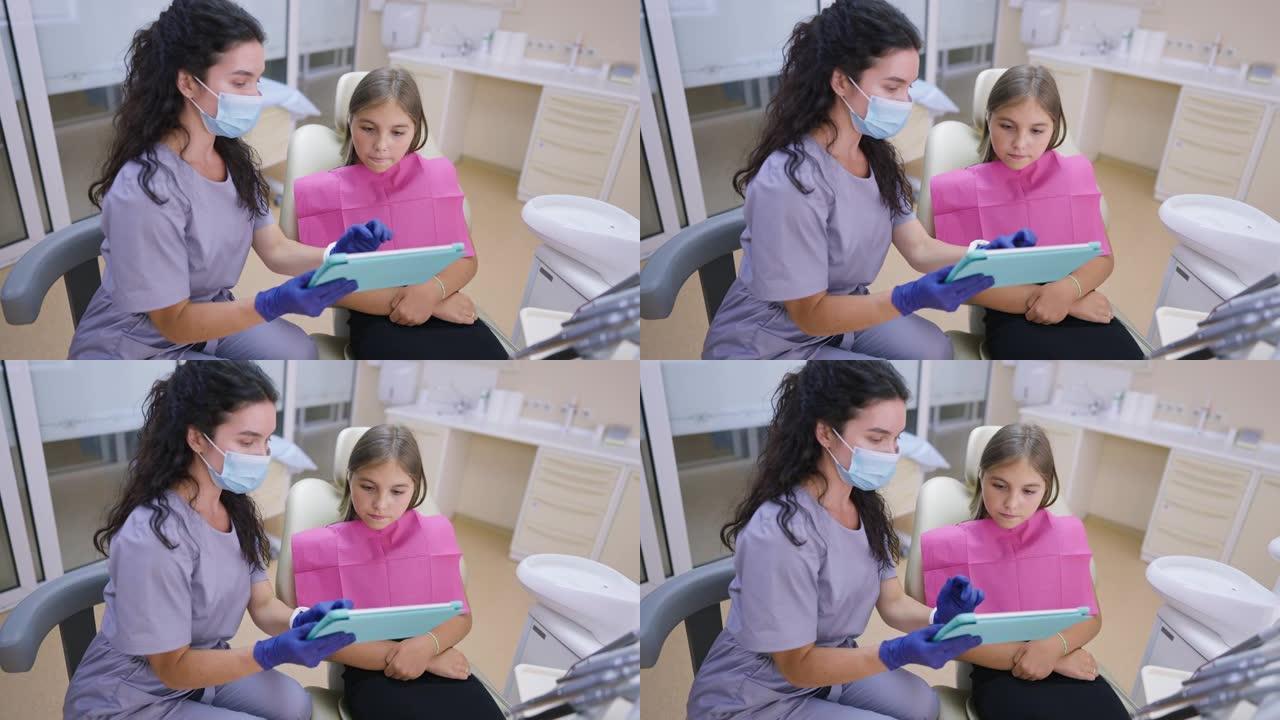 可爱的古玩少女坐在牙医椅上听专业医生展示平板电脑说话。感兴趣的高加索少年在室内医院探望正畸医生的肖像