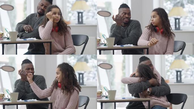 非洲裔美国父亲与十几岁的女儿握手拥抱和微笑。正面英俊的男人和可爱的少女在室内坐在家里的桌子上发誓。