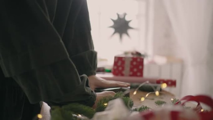 组装和准备圣诞礼物的侧视图。一个女人的特写镜头用带有圣诞节装饰品的包装纸将她的手包裹在盒子上。专业装