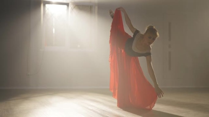 自信的高加索女芭蕾舞演员，一条腿向前弯曲并旋转。美丽苗条芭蕾舞演员的肖像在舞蹈工作室的背光雾中排练表