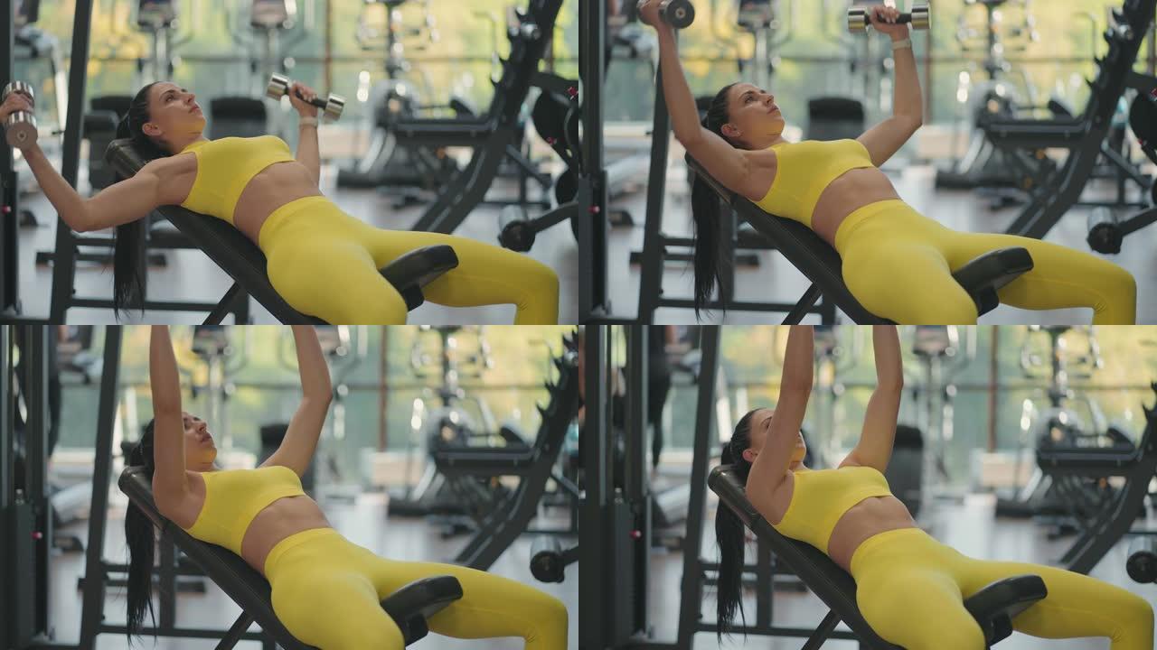英俊的运动女子躺在健身房里通过繁殖哑铃来训练胸肌。肌肉发达的女运动员用哑铃锻炼，举重