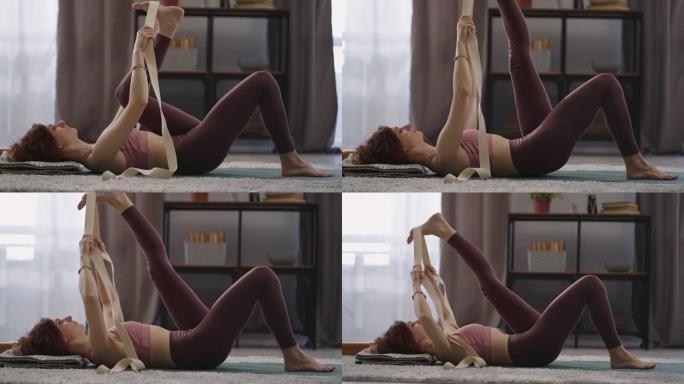 女人在家做伸展运动，躺在地板上，用胶带拉伸腿部肌肉，身体状况良好