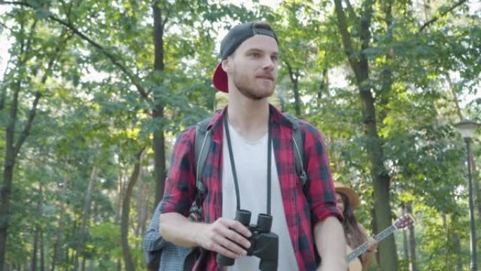 充满信心的男性向导在夏季森林中，一群积极的年轻游客在后台漫步。英俊的白人男子走路和离开的肖像，微笑着