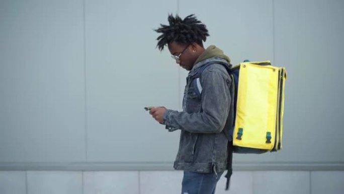英俊的非洲裔美国男性快递员使用智能手机带着黄色保温袋在城市行走的侧视图。寻找送货地址的年轻人。服务和