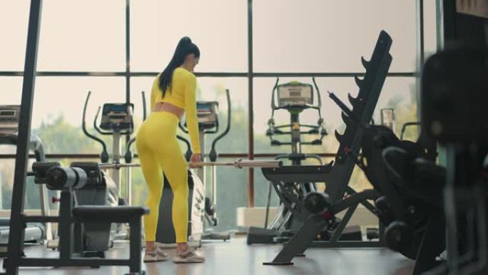 适合西班牙裔女性穿着黄色运动服在健身房用杠铃进行举重硬拉运动。女人黑发健身表演和杠铃做硬拉运动