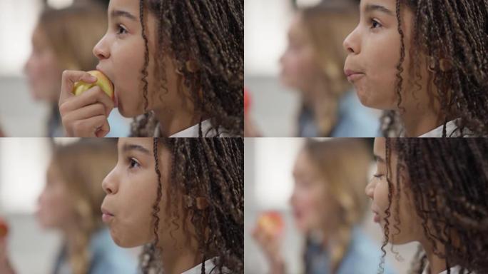 快乐美丽的非洲裔美国女孩吃健康苹果的侧视图特写，背景是模糊的高加索朋友。脸积极迷人的孩子享受健康的食