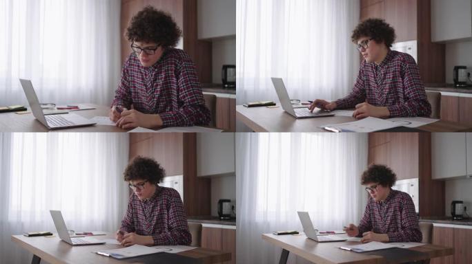 卷发男学生戴眼镜的迷人小男孩正在家里学习，用笔记本电脑打字，用笔记本电脑写字。大学生使用笔记本电脑观