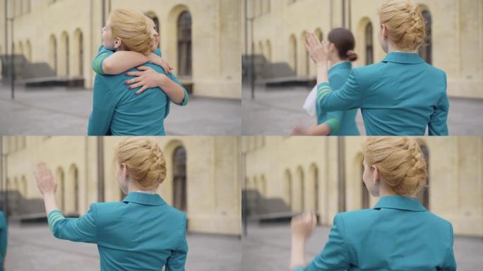 年轻金发空姐拥抱模糊的黑发同事，挥手告别。城市街道上苗条自信的白人同事道别。职业和美丽。