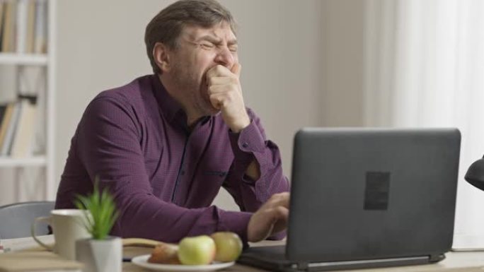 无聊的昏昏欲睡的人在笔记本电脑键盘上打字，打哈欠和伸展坐在室内的桌子上的中间镜头。疲惫的高加索中年男
