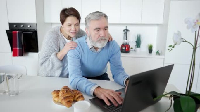 忙碌的白人男子在笔记本电脑上在线发送消息的肖像，是充满爱心的女人拥抱丈夫微笑和交谈。幸福的退休夫妇在
