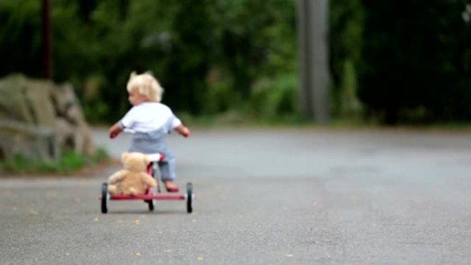 可爱的小男孩，带着泰迪熊玩具，在雨中骑着三轮车在街上，赤脚，夏天