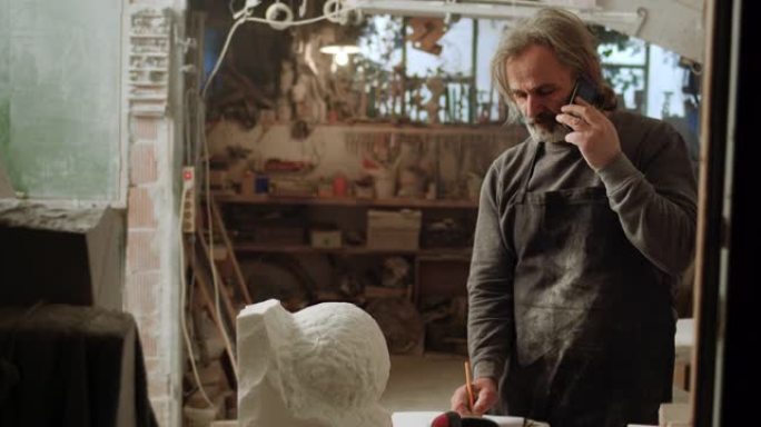SLO MO男雕塑家在工作室里打电话并做笔记