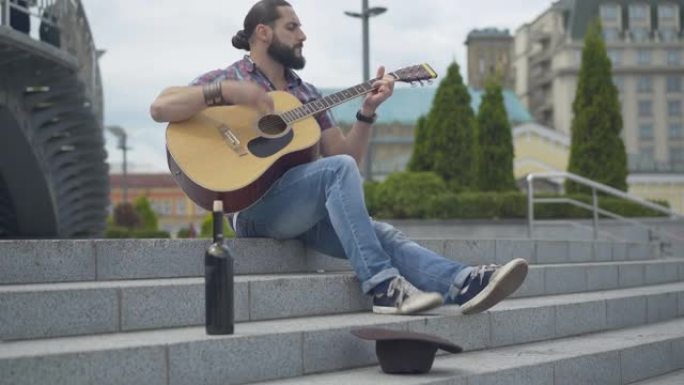广角拍摄的肖像热情的音乐家弹奏吉他在城市的楼梯与酒瓶和帽子躺在前面。可怜的白人在户外表演赚钱。贫困和
