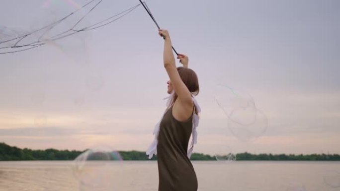 一个年轻的嬉皮士女人穿着裙子，头上长着羽毛，在日落时在湖岸慢动作时产生巨大的肥皂泡