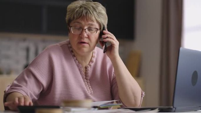 老年女性专家在冠状病毒大流行期间远程工作，女性正在使用笔记本电脑和计算器，通过电话拨打