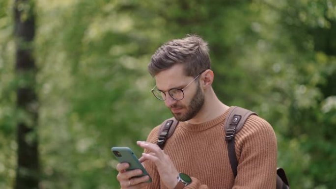 男子徒步旅行者在森林里享受周末时使用智能手机。手机上迷人的男人背包客规划森林旅行路线的低角度视图