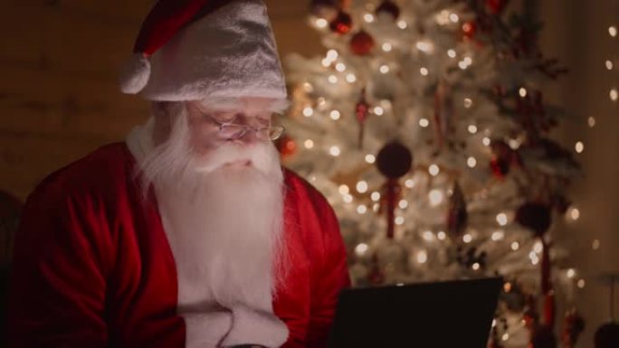 圣诞老人晚上在圣诞树背景上的圣诞灯的灯光下用笔记本电脑工作