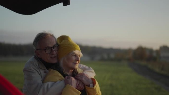 两个老年人在公路旅行中露营。站在汽车旁边拥抱。相机平移到绿色领域