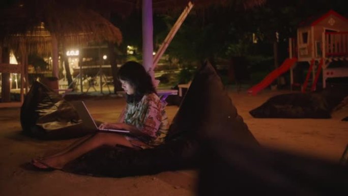 年轻的亚洲妇女在键盘上打字，晚上在海边度假享受时使用笔记本电脑触控板。