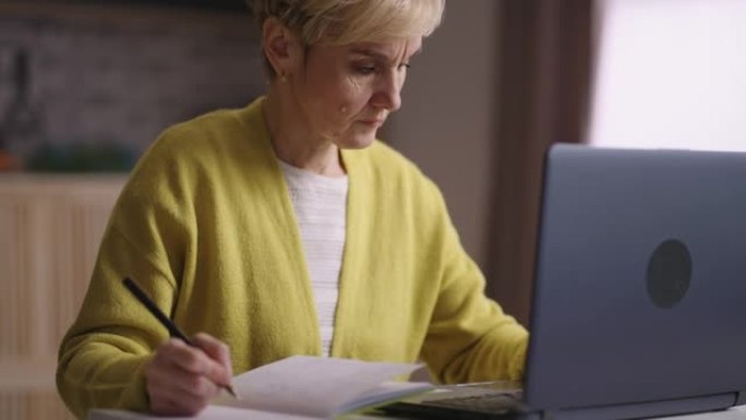老太太正在网上学习或工作，在笔记本电脑上上网，在笔记本电脑上做笔记，在家中的退休妇女肖像