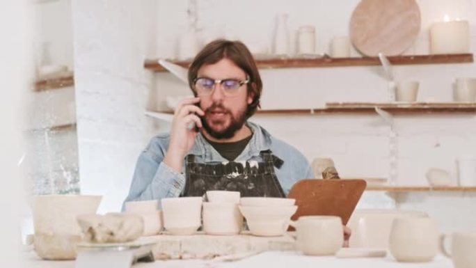 陶器艺术家在陶瓷艺术工作室里打电话和使用剪贴板。艺术创意陶工在业余工艺车间中使用技术进行网络交流，就