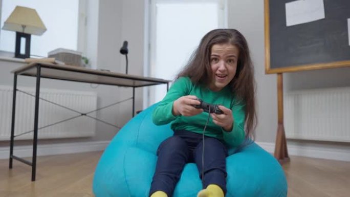 女性玩家坐在室内的书包椅子上，在在线视频游戏中欢欣鼓舞。兴奋的高加索小人物的肖像使用游戏控制器看着相