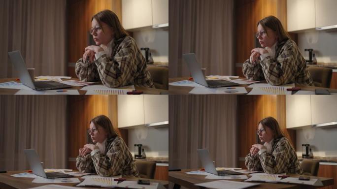 一位戴着眼镜的沉思女人看着笔记本电脑的屏幕，集中精力，并提出了发展策略。纸上的时间表和计划已经摆在桌
