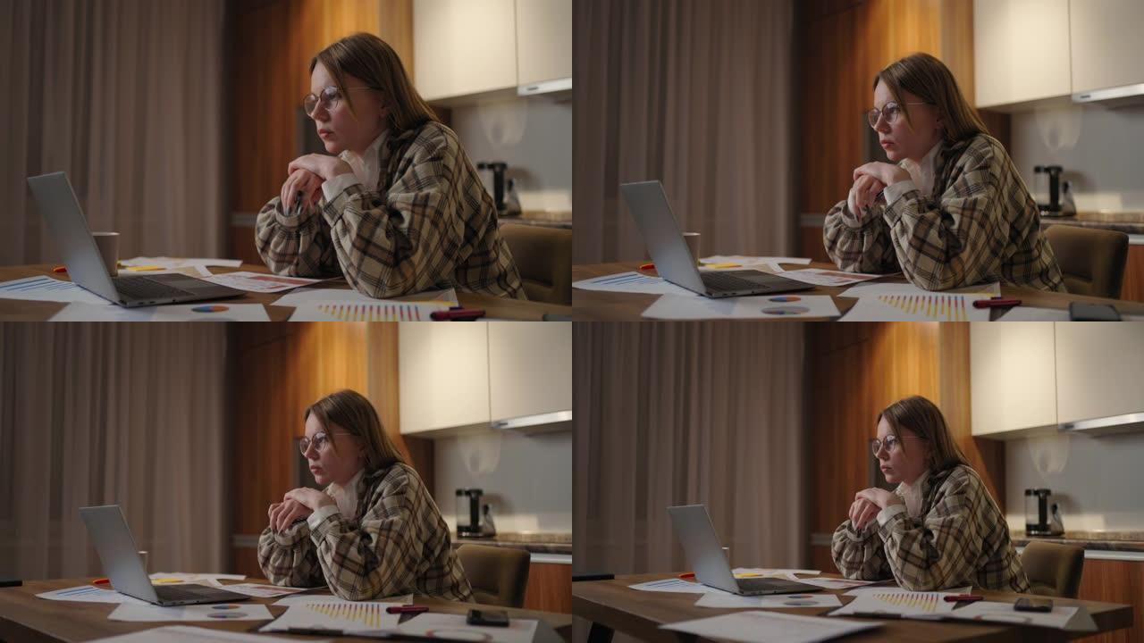 一位戴着眼镜的沉思女人看着笔记本电脑的屏幕，集中精力，并提出了发展策略。纸上的时间表和计划已经摆在桌
