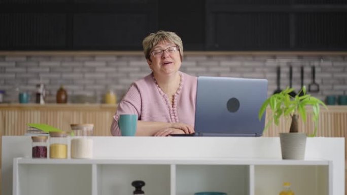 快乐的老年妇女通过笔记本电脑上的视频聊天进行交流，退休女士周末坐在家里的厨房里