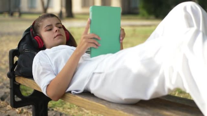 放松积极的年轻女子躺在大学院子的长凳上，戴着耳机听音乐。自信美丽苗条的白人学生在户外休息时享受爱好的