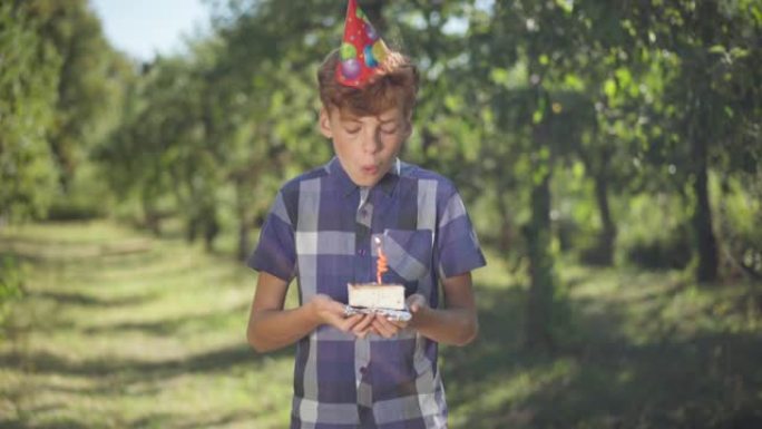 积极的红发少年在生日蛋糕上吹蜡烛站在春夏公园的阳光中摆姿势。在户外阳光明媚的日子里，戴着党帽的快乐少