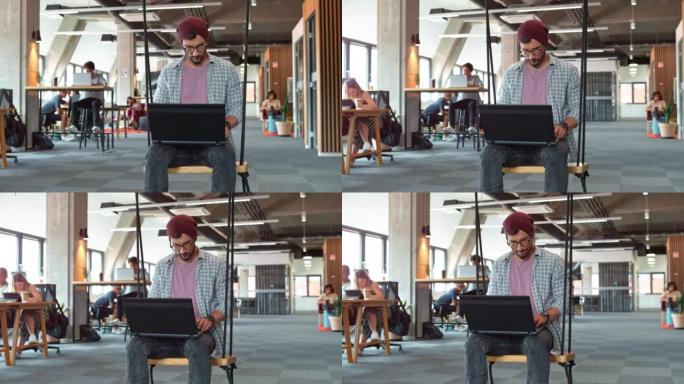 戴无檐小便帽的年轻人坐在办公办公室的秋千上，在笔记本电脑上工作