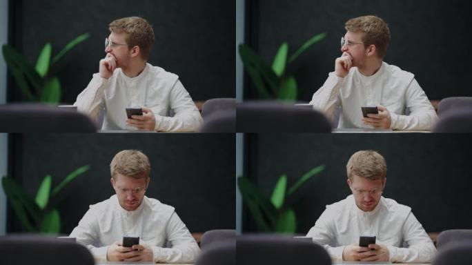 戴着眼镜和白衬衫的英俊沉思的男人独自坐在咖啡馆里，手里拿着智能手机