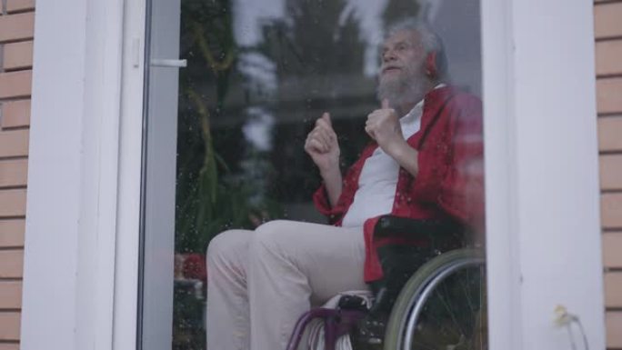 坐在轮椅上的开朗的大胡子残疾老人戴着耳机欣赏音乐，模仿鼓演奏。室内玻璃门后积极快乐的高加索残疾人退休