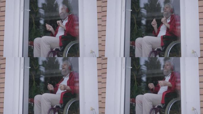 坐在轮椅上的开朗的大胡子残疾老人戴着耳机欣赏音乐，模仿鼓演奏。室内玻璃门后积极快乐的高加索残疾人退休