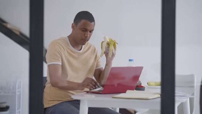 用严肃的非裔美国人的杯子射击，他在笔记本电脑键盘上用香蕉打字，表情集中。专注的年轻经理在家庭办公室在