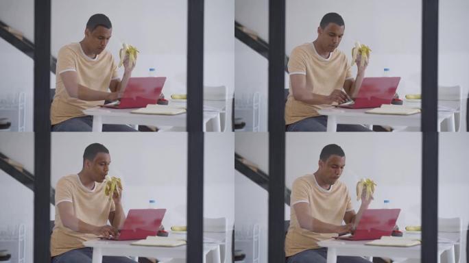 用严肃的非裔美国人的杯子射击，他在笔记本电脑键盘上用香蕉打字，表情集中。专注的年轻经理在家庭办公室在