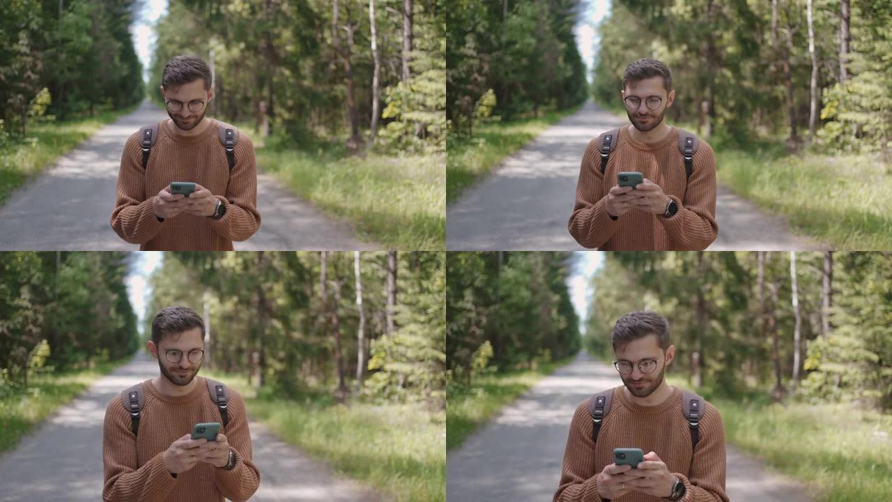 一个背着背包的年轻人沿着森林中的一条土路走，写信息，在互联网上交流，看内容，微笑。