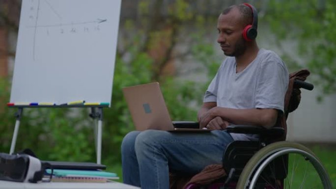 严肃的非洲裔美国职业男子戴着耳机的侧视图在笔记本电脑上的视频聊天中慢动作交谈。自信的残疾瘫痪户外商业