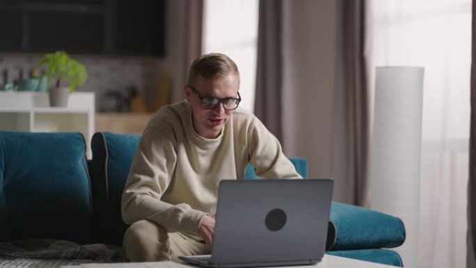 男子坐在沙发上在家使用笔记本电脑进行视频隔离。快乐的年轻金发老师在家工作，在笔记本电脑视频会议上聊天
