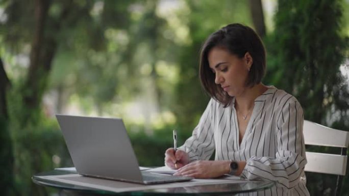 学生女子布鲁内特阿拉伯西班牙裔民族通过互联网远程学习，坐在夏季咖啡馆里拿着笔记本电脑，写下笔和笔记本