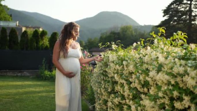 穿着白色连衣裙的孕妇站在花丛中的花园里，抱着肚子，看着风景