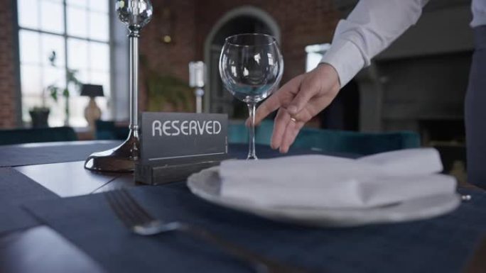 男性手将酒杯放在桌子上，并带有预留标志。无法辨认的高加索服务员在室内慢动作的豪华餐厅摆台。