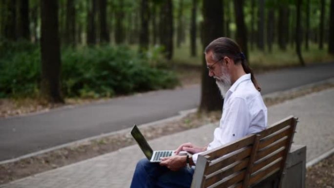 一个成年男子在公园长椅上留着灰色胡须，与笔记本电脑一起工作。4k慢动作视频。